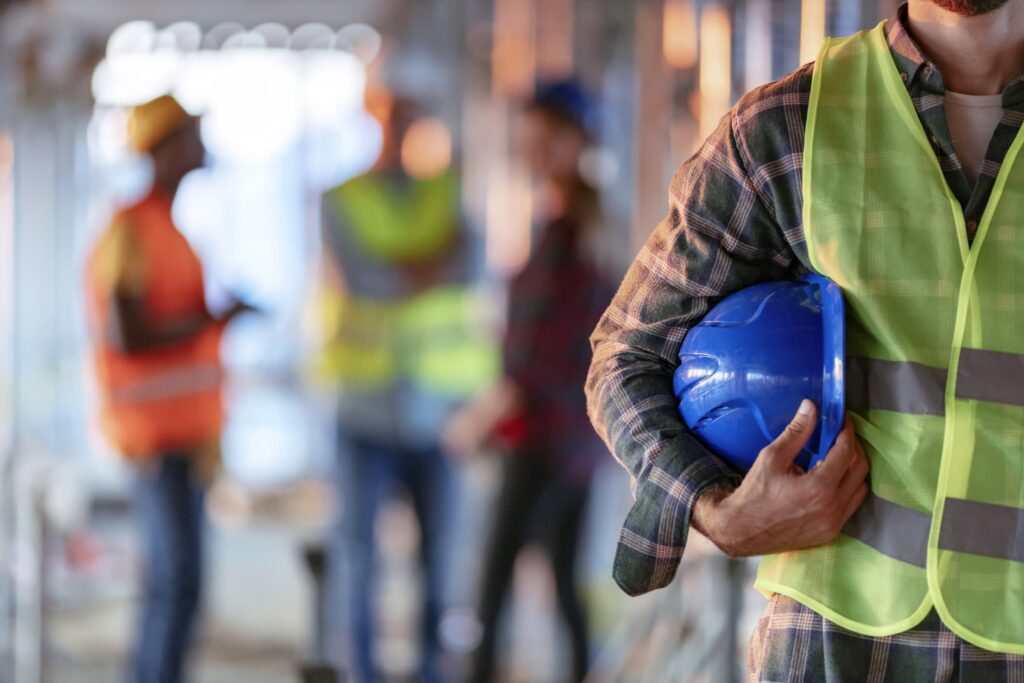 Close up of man holding work helmet there are more tradesmen standing in the background