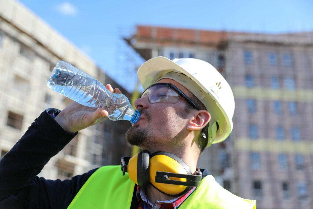 Male tradesman working outdoors in high-vis vest and protective gear drinking bottle of water to protect himself from heat injury