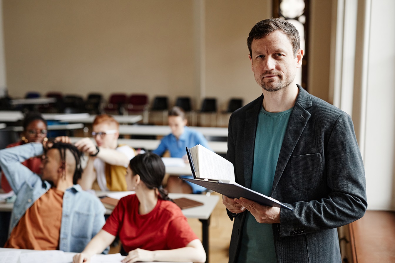 Highschool teacher holding book standing in classroom with students behind him